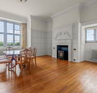 A bright dining room with large windows, wooden floor, six chairs around a glass table, a fireplace, and light-colored walls with wainscoting.