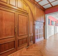 A corridor with wooden paneling and flooring, red walls with framed pictures, and white wainscoting. A large wooden door is on the left, and the hallway extends into another room.