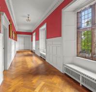 A long hallway with white wainscoting and red walls features a large leaded-glass window and framed artwork. The space is illuminated by natural light.