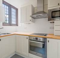 A modern kitchen with wooden countertops, a metal sink, an electric stovetop, an oven, a microwave, and white cabinetry. A window with a wooden frame is above the sink.