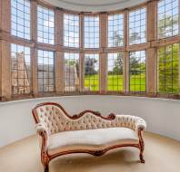 A white, tufted chaise lounge with a wooden frame is placed in a circular room surrounded by large, arched windows overlooking a lush, green landscape.