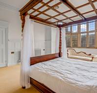 A bedroom with a four-poster bed, sheer curtains, and a tufted chaise lounge by large, multi-pane windows. White walls and carpeted floor are visible.