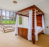A bedroom with a wooden four-poster canopy bed, sheer white curtains, a curved vintage chaise lounge, and large bay windows overlooking a green landscape.