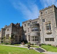 A large historic stone mansion with multiple windows and turrets, surrounded by a well-maintained lawn and garden, set against a clear blue sky.