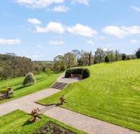 A landscaped garden with manicured lawns, stone pathways, and wooden benches surrounded by trees and hills on a clear day with a blue sky and white clouds.