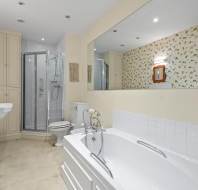 A bathroom with a bathtub, sink, toilet, and a shower enclosed in glass. The walls have floral wallpaper and there is a large mirror above the sink. The floor is tiled.