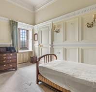 A small bedroom with cream-colored paneled walls, a single bed, a chest of drawers, a window with green curtains, a wall-mounted lamp, and a painting above the dresser.