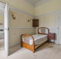 A bedroom with two single beds, one closer to the camera and another in an adjacent room, featuring light-colored walls, a chest of drawers, wall lamps, wooden doors, and beige carpet.