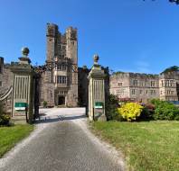 A historic stone castle with two tall towers, an arched entrance, and a gravel driveway, surrounded by manicured gardens and lawns under a clear blue sky.