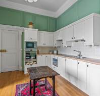 A kitchen with green walls, white cabinets, wooden countertops, and a wooden floor. There is a small wooden table on a red patterned rug in the center. Appliances are integrated into the cabinetry.