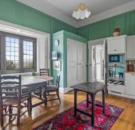 A traditional kitchen with green walls and white cabinets, featuring wooden chairs around a round table, a small central table, and a red rug on wooden flooring, with large windows and built-in storage.