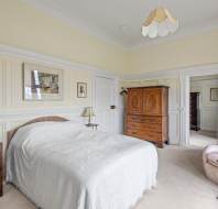 A bedroom with a double bed, white bedspread, two wooden nightstands, a wooden dresser, a closet, and a peach armchair. Walls are paneled in light colors, and there is a framed picture above the bed.