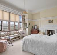 A bedroom with a large window, floral curtains, an armchair, a bed with a patterned white cover, a fireplace, and various pieces of furniture including a dresser and a bench.
