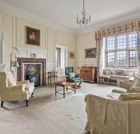 A traditional living room with vintage furniture, a fireplace, large windows, and framed portraits. The room has neutral tones with white walls, floral curtains, and a carpeted floor.