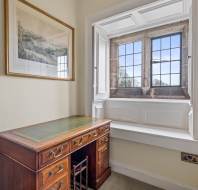A small study nook with a wooden desk, green leather top, and a chair. There is a framed painting on the wall and a large window with a view outside. Shelves are partially visible on the right.