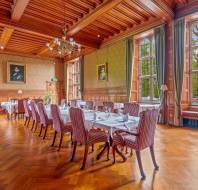 A refined dining room with large windows, wood-paneled walls, a parquet floor, striped upholstered chairs around set tables, and chandeliers.