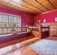 A Victorian-style room with wooden paneling, a large stained glass window, cushioned benches by the window, a chandelier hanging from the wooden ceiling, and framed pictures on the wall.