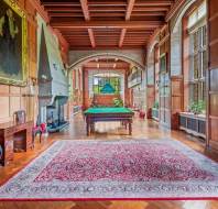 A spacious, wood-paneled room with a billiards table, ornate rug, large windows, a fireplace, and elegant furniture, including a desk and chairs, under a coffered ceiling.