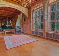Spacious room with large windows, wooden ceiling, ornate rug, and a billiards table. Several potted plants and armchairs are placed around the room, with a staircase leading to the next level.