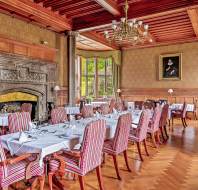 Elegant dining room with striped upholstered chairs, neatly set tables with white tablecloths, a large ornate fireplace, wooden paneling, and a chandelier. A portrait hangs on the wall near windows.