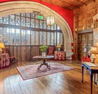 A grand, historical room with a large wooden arch, stained glass windows, a central round table with a plant, and two side tables with lamps on either side, adorned with red and gold decor.