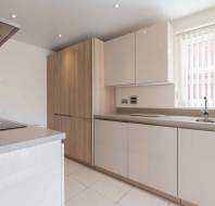 Modern kitchen with light wood and white cabinets, a sink under a window with vertical blinds, and built-in appliances on a light tiled floor.