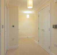 A hallway with beige walls and carpet, featuring closed white doors, a light fixture on the ceiling, and various wall switches and outlets.