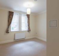 Empty room with beige walls and carpet, two windows with blinds and patterned curtains, ceiling light, and a radiator beneath the windows.
