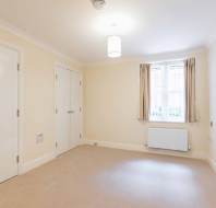 Empty beige carpeted room with cream walls, a ceiling light, two closed white doors, a window with beige curtains, and a white radiator beneath the window.