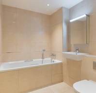 Modern bathroom with beige tiled walls, bathtub with handheld shower, wall-mounted sink, toilet, towel radiator, and mirrored cabinet under bright lighting.