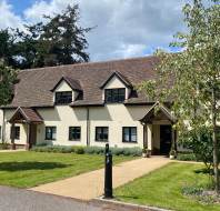 A two-story cream-colored building with a steep tiled roof, black window frames, and two entrances, surrounded by grass, trees, and a pathway under a partly cloudy sky.