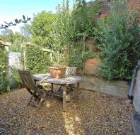 A small outdoor patio with a round wooden table, two chairs, and potted plants, surrounded by greenery and a brick wall, on a gravel surface.