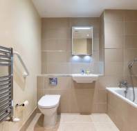 A modern bathroom with beige tiles, featuring a wall-mounted toilet, a sink, a mirror with lights, a bathtub with a showerhead, and a metallic towel rack.