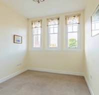 A small, empty room with beige walls, three windows with patterned curtains, carpeted floor, simple ceiling light, and a framed picture on one wall.