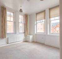 Bright, empty room with beige carpet, white walls, two large windows with blinds, a small chest of drawers, and a radiator beneath the windows.