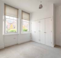 An empty room with beige carpet, two large windows with blinds, white walls, a white ceiling, and closed double-door closets. There's a small white dresser on the left.