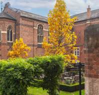 A brick church with arched windows is surrounded by vibrant autumn trees and greenery. A small signpost is visible in the garden area.