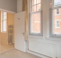 A room with beige walls, a radiator, and tall windows. An open door reveals a bathroom with beige tiles and a towel warmer. Beige curtains hang beside the windows.