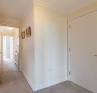 A corridor with cream-colored walls, two closed doors, framed pictures on the wall, and a view into a sunlit room.