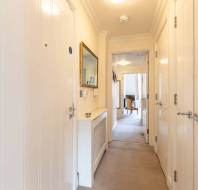 A narrow hallway with white paneled doors, a wall-mounted radiator, and a mirror. The hallway leads to a room with a chair and visible natural light.