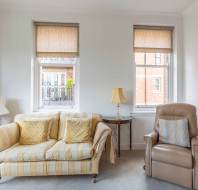 A cozy living room with a striped sofa, cushioned chairs, two windows with blinds, and side tables with lamps.