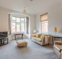 A bright living room with a bay window, beige sofa, armchair, ottoman, TV on a stand, and two side tables with lamps.