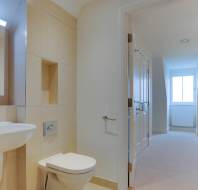 A clean, modern bathroom with a wall-mounted sink and toilet, beige tiles, and an open door leading to a bright room with carpet.