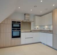 Modern kitchen with light wooden cabinets, white countertops, built-in oven, stovetop, and stainless steel cooker hood. Recessed lighting and tiled floor.