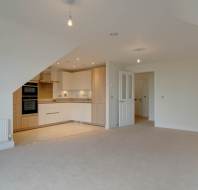 Empty room with beige carpet, an open kitchen with wooden cabinets, white walls, recessed lighting, and two ceiling bulbs. A white door leads to another room.