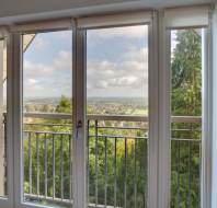 View through large glass doors of a balcony with a railing, overlooking a landscape with trees and distant fields under a partly cloudy sky.