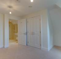 A carpeted empty room with open double doors leading to a small bathroom. The walls are painted white, and there are ceiling lights in both areas.