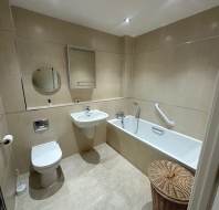 A clean beige-tiled bathroom featuring a toilet, sink, bathtub with grab bar, a wall mirror, and a wicker laundry basket.