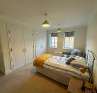 A bedroom with a double bed, yellow blanket, cushions, and bedside lamp. White wardrobes line one wall, and there are two windows with patterned blinds and a gray chair underneath.