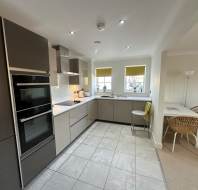 Modern kitchen with grey cabinets, white countertops, built-in oven, and stove. Two windows with yellow blinds, a small dining table with chairs, and tiled floor.